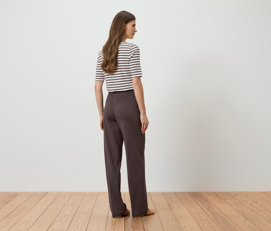 Une femme portant un pantalon marron et un haut rayé se tient devant un mur blanc.