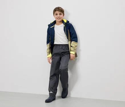 Un garçon souriant en veste de pluie, pantalon et bottes est appuyé contre un mur.