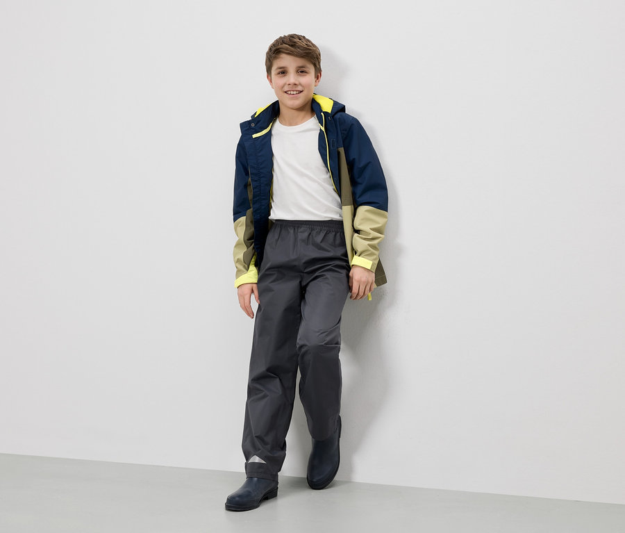Un garçon souriant en veste de pluie, pantalon et bottes est appuyé contre un mur.