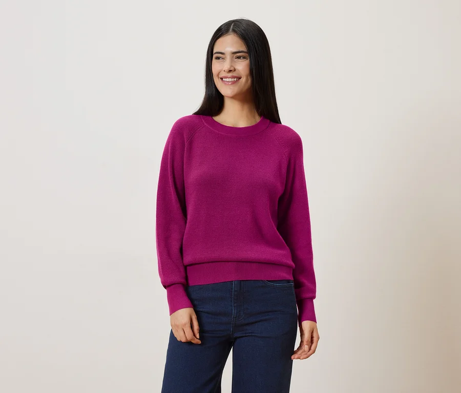 Une femme aux longs cheveux foncés porte un pull rose et un jean bleu sur un fond clair.