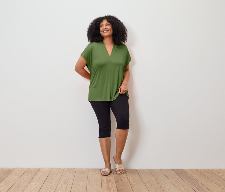 Une femme aux cheveux bouclés porte un t-shirt vert, un pantacourt noir et des sandales devant un mur blanc.