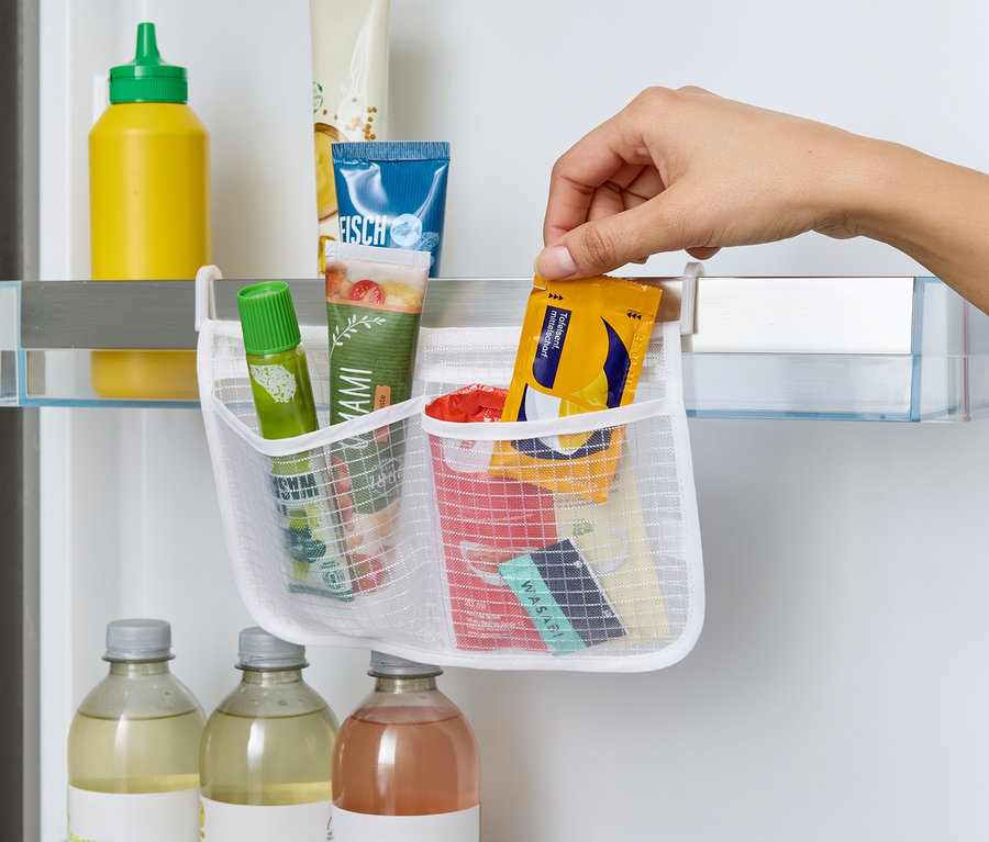 Une main place un paquet jaune dans un organisateur en filet blanc accroché à une étagère de réfrigérateur. À l'intérieur, tubes, sauces et wasabi.