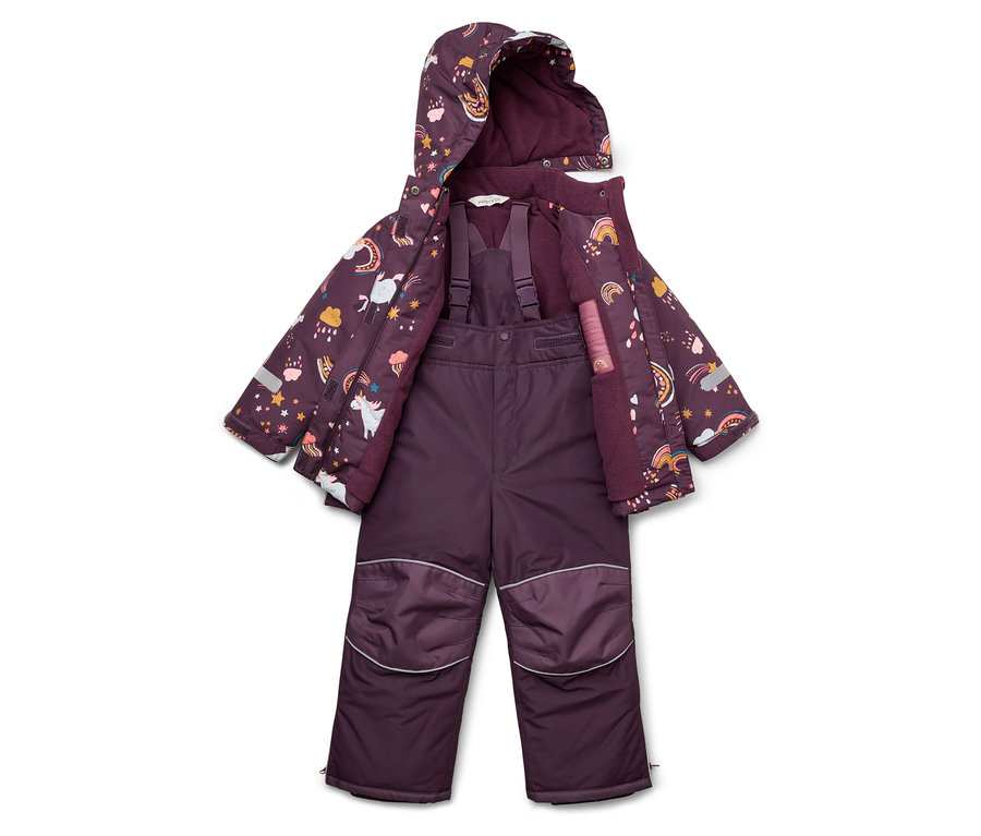 Une combinaison de ski violette pour enfant et une veste à motifs de licornes, d'étoiles et d'arcs-en-ciel sont posées sur le sol.
