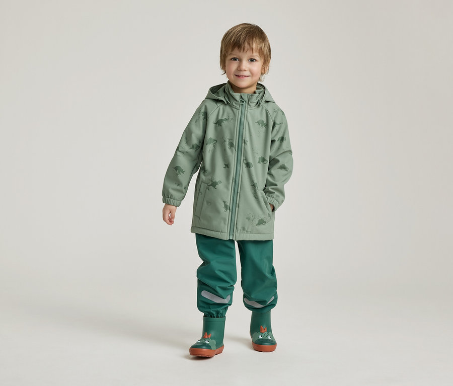 Un garçon se tient sur un fond clair, vêtu d'un manteau de pluie vert à motifs de dinosaures, d'un pantalon vert et de bottes en caoutchouc.
