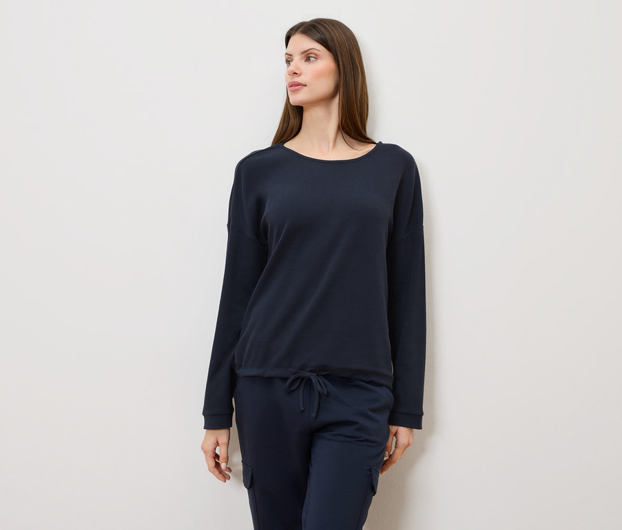 Femme aux longs cheveux bruns portant un pull bleu marine et un pantalon bleu marine regardant sur le côté.