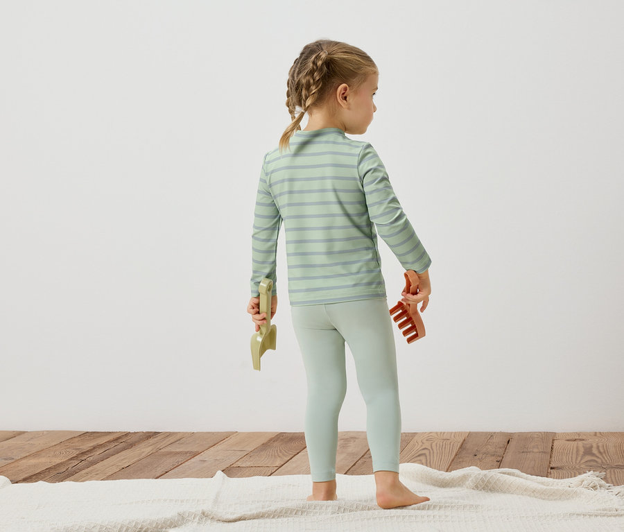 Une jeune fille, vue de dos, porte un haut rayé et un legging, et tient des jouets dans ses mains.