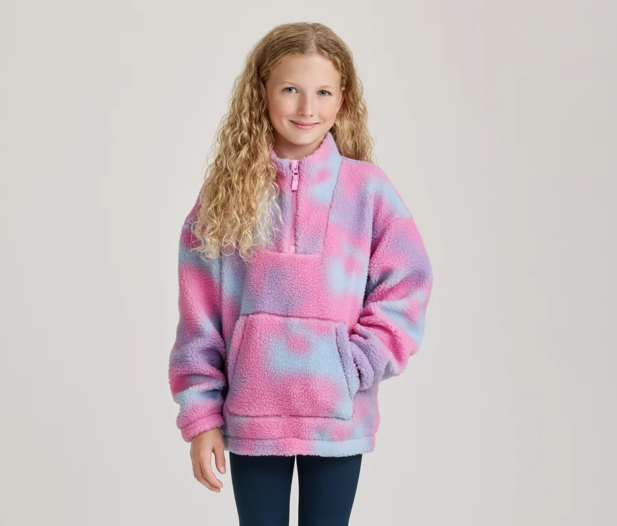 Jeune fille souriante aux cheveux blonds bouclés, portant un pull polaire tie-dye rose, violet et bleu avec un demi-zip et une poche.
