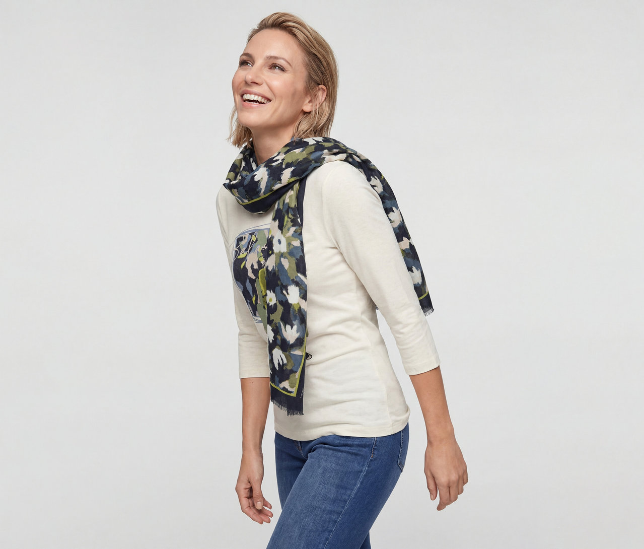 Femme souriante aux cheveux blonds courts portant un haut crème à manches longues, un jean bleu et une écharpe à motifs.