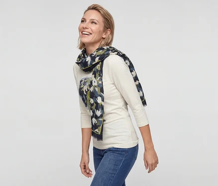 Femme souriante aux cheveux blonds courts portant un haut crème à manches longues, un jean bleu et une écharpe à motifs.