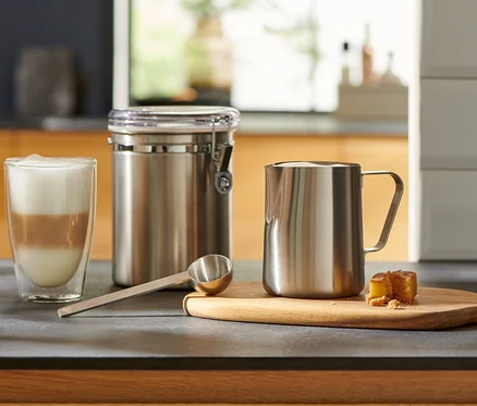 Sur le comptoir de la cuisine, il y a un verre de café latte, une cuillère doseuse argentée, un récipient et un pot à lait.