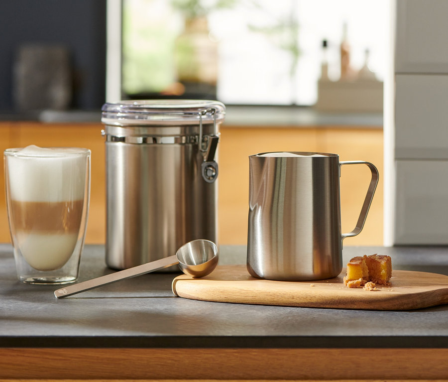 Sur le comptoir de la cuisine, il y a un verre de café latte, une cuillère doseuse argentée, un récipient et un pot à lait.