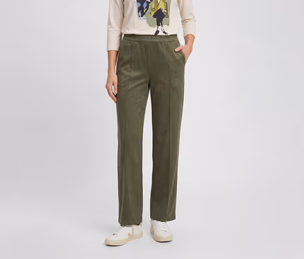 Une personne porte un pantalon droit vert olive à taille élastique, un t-shirt clair à manches longues imprimé et des baskets blanches. Une main est dans la poche.