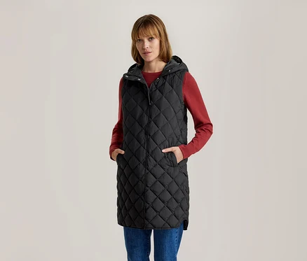 Femme en longue veste matelassée noire à capuche, t-shirt rouge à manches longues et jean bleu, mains dans les poches.