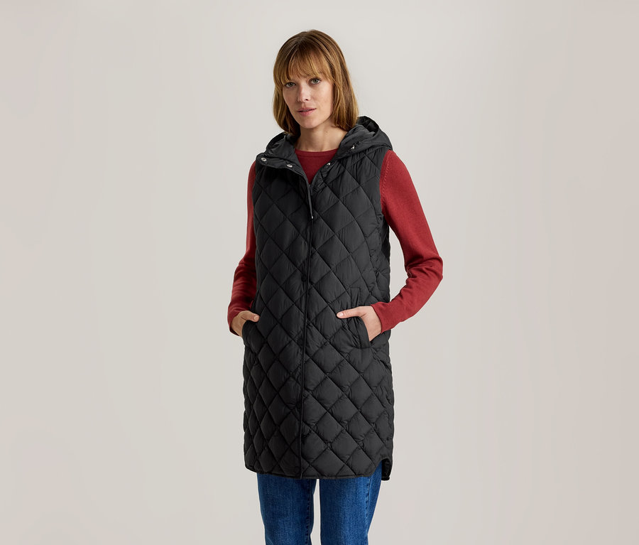 Femme en longue veste matelassée noire à capuche, t-shirt rouge à manches longues et jean bleu, mains dans les poches.