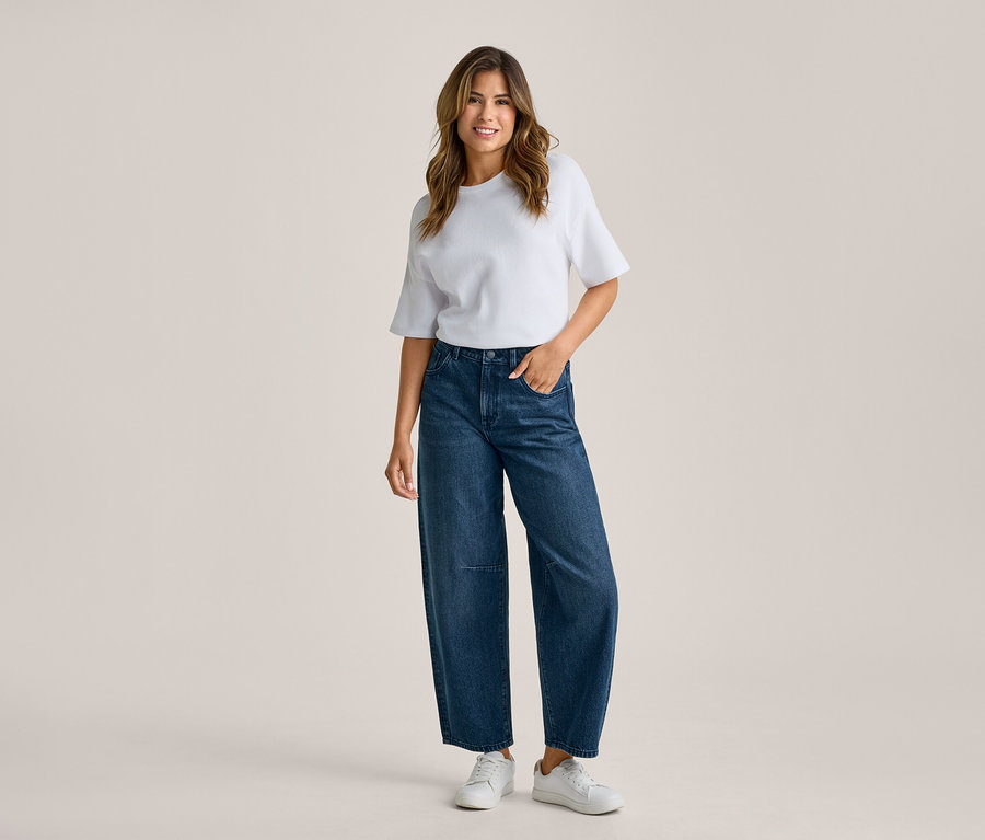 Femme debout, souriante, portant un t-shirt blanc, un jean large bleu foncé et des baskets blanches, une main dans la poche.