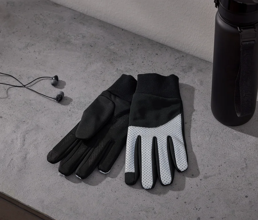Deux gants noirs et blancs avec des paumes pointillées, des écouteurs filaires noirs et une bouteille d'eau noire sur une surface en béton gris.