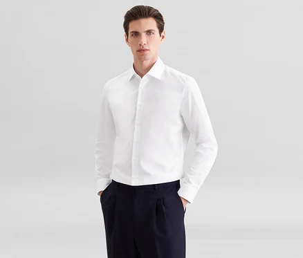 Homme portant une chemise blanche à manches longues et un pantalon plissé bleu foncé, les mains dans les poches.