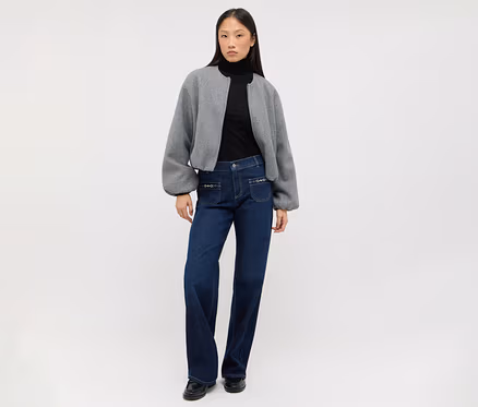 Femme portant un col roulé noir, une veste courte grise à manches bouffantes, un jean large bleu foncé avec des poches décoratives et des chaussures plates noires.