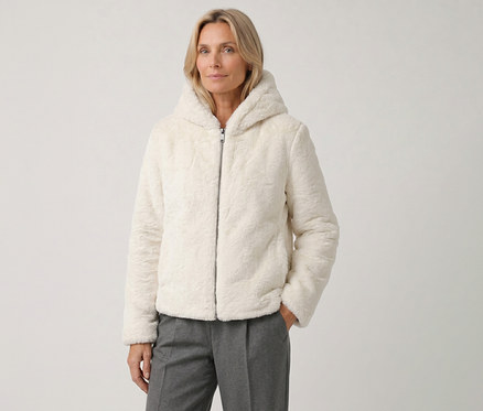 Une femme blonde porte une veste à capuche moelleuse de couleur crème et un pantalon gris, sa main droite dans la poche.