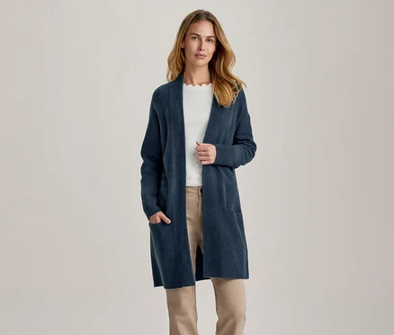 Femme portant un long cardigan bleu foncé ouvert sur un haut blanc et un pantalon beige, une main dans la poche.