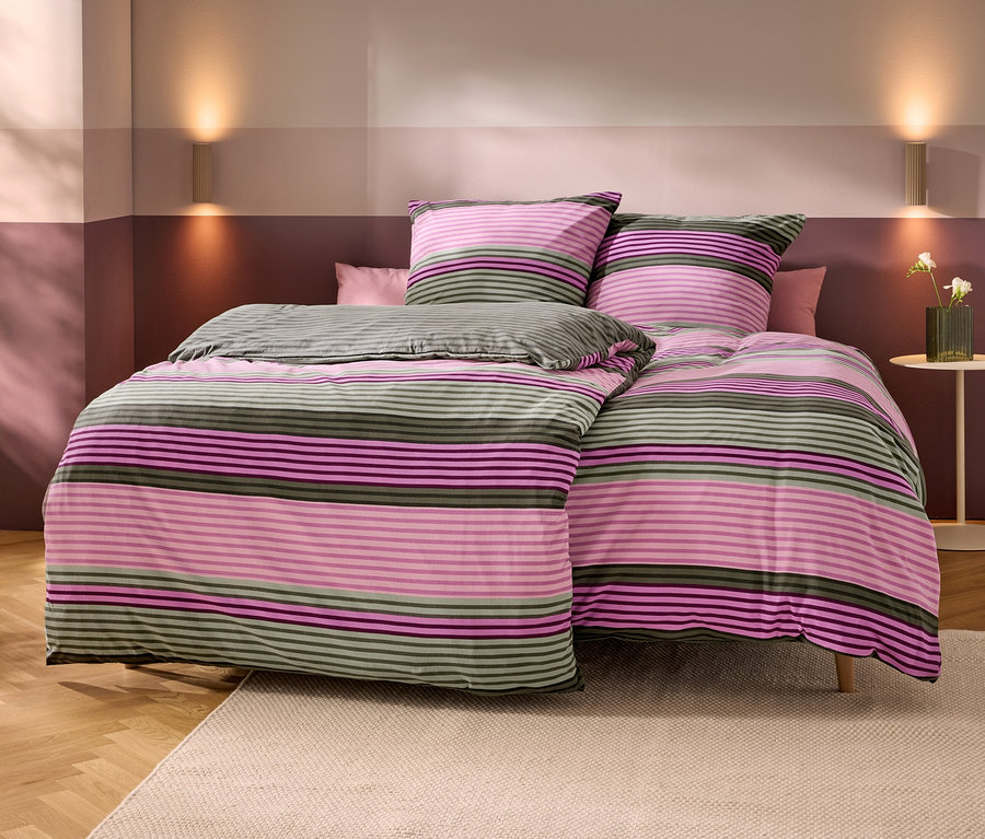 Un lit avec une parure de lit rayée rose, violette et vert olive. Deux oreillers assortis et un petit oreiller rose uni sont posés dessus. Les murs sont bicolores en rose clair et foncé/violet, avec deux appliques murales. À droite, une table d'appoint avec un vase de fleurs blanches. Plancher en bois avec un tapis beige.