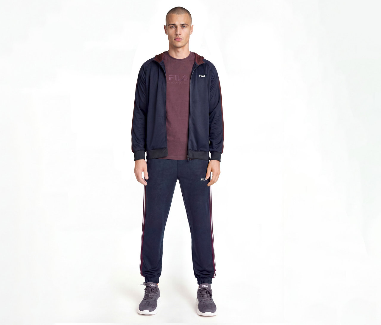 Homme portant un survêtement Fila bleu marine avec des rayures latérales bordeaux, un t-shirt bordeaux et des baskets grises, debout devant un fond blanc.