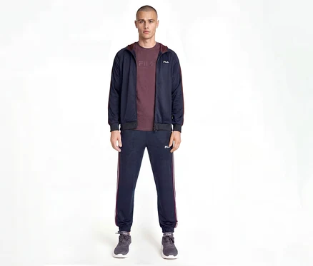 Homme portant un survêtement Fila bleu marine avec des rayures latérales bordeaux, un t-shirt bordeaux et des baskets grises, debout devant un fond blanc.