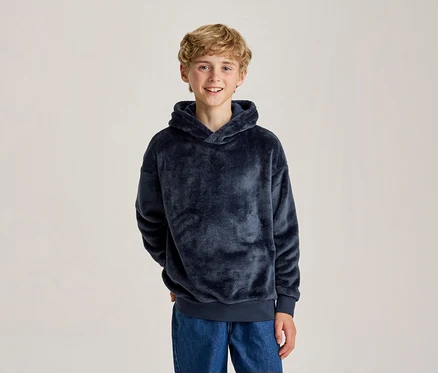 Jeune garçon souriant aux cheveux blonds bouclés, portant un sweat à capuche bleu foncé moelleux et un jean bleu, les mains derrière le dos.