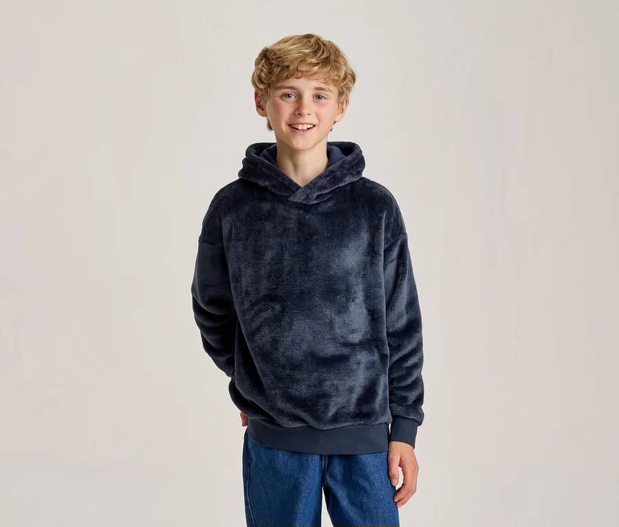 Jeune garçon souriant aux cheveux blonds bouclés, portant un sweat à capuche bleu foncé moelleux et un jean bleu, les mains derrière le dos.