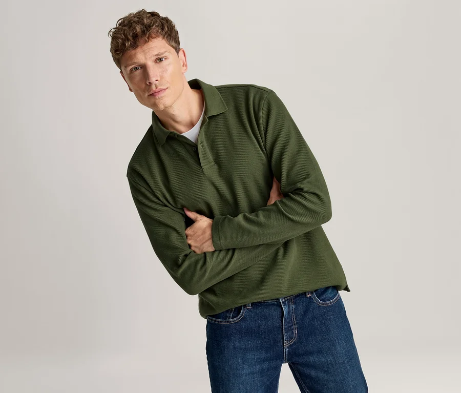 Homme aux cheveux bruns bouclés portant un polo vert foncé à manches longues et un jean bleu foncé, les bras croisés, regardant la caméra.
