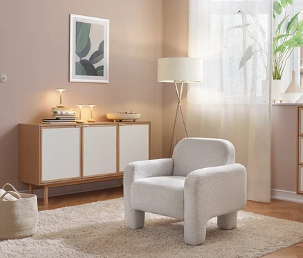 Une pièce confortable avec un fauteuil gris clair sur un tapis moelleux. À gauche, un buffet en bois clair avec des portes blanches, surmonté de lampes et de livres. À droite, un lampadaire et une commode près d'une fenêtre avec une plante.