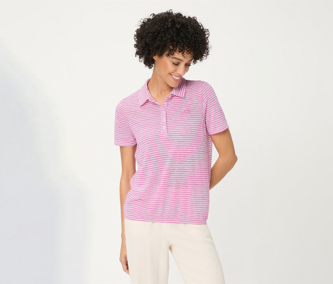 Femme souriante aux cheveux bouclés portant un Polo Olsen rose rayé et un pantalon clair, regardant vers le bas.