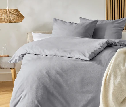 Un lit avec une parure de lit gris clair et deux oreillers, un plaid crème à franges. À gauche, un tabouret avec une carafe en verre, au-dessus duquel pend une lampe tressée. Le sol est en bois, avec un tapis clair devant.