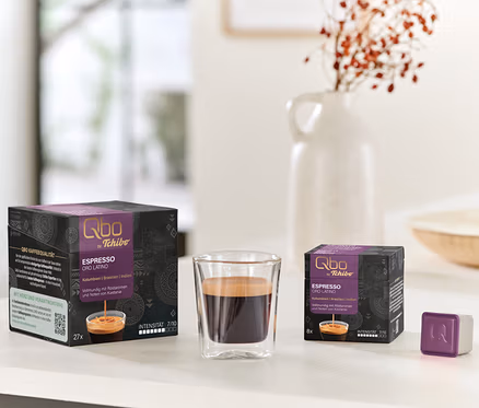 Sur une table, des paquets de capsules Qbo Espresso ORO TOLIMA (27 et 8 capsules) sont posés à côté d'un verre d'espresso et d'une capsule individuelle.