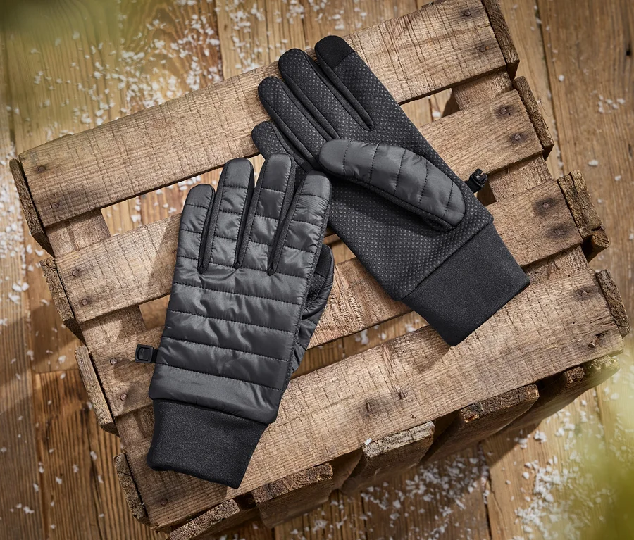 Deux gants noirs, l'un avec un dos matelassé et l'autre avec une paume texturée, sont posés sur une caisse en bois avec des flocons blancs éparpillés.