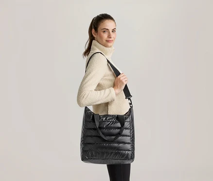 Une femme aux cheveux attachés en queue de cheval, vêtue d'un pull polaire col roulé crème et d'un legging noir, porte un sac fourre-tout matelassé noir à l'épaule et regarde l'objectif en souriant légèrement.
