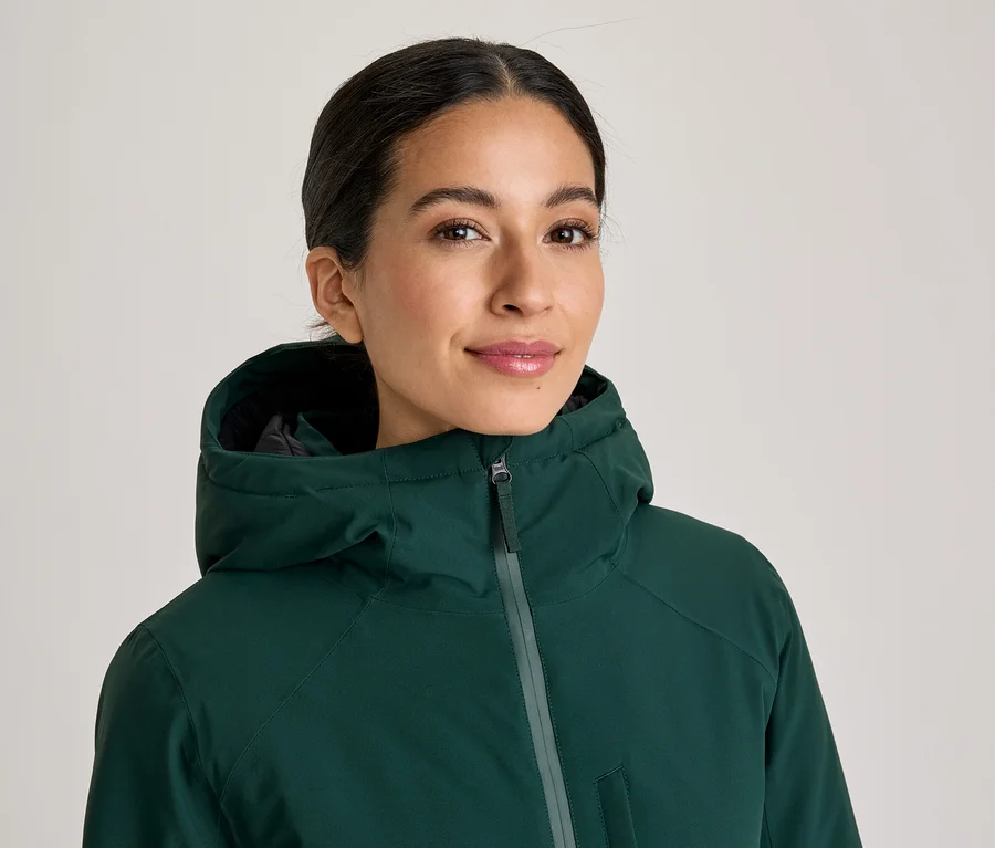 Femme souriante portant une veste à capuche vert foncé, regardant directement l'appareil photo.