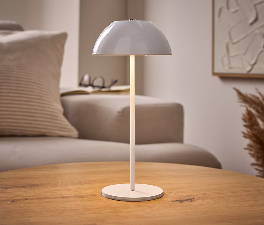 Lampe de table à LED sans fil blanche allumée sur une table en bois, avec canapé beige, livre et art abstrait en arrière-plan.