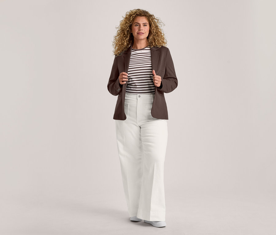 Une femme aux cheveux bouclés se tient de face, tenant les revers de son blazer marron ouvert. Elle porte un t-shirt rayé noir et blanc, un pantalon blanc à jambes larges et des chaussures plates de couleur claire.