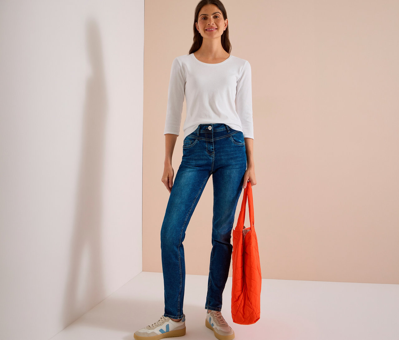 Femme souriante portant un haut blanc à manches longues et un jean CECIL bleu, tenant un sac cabas orange.