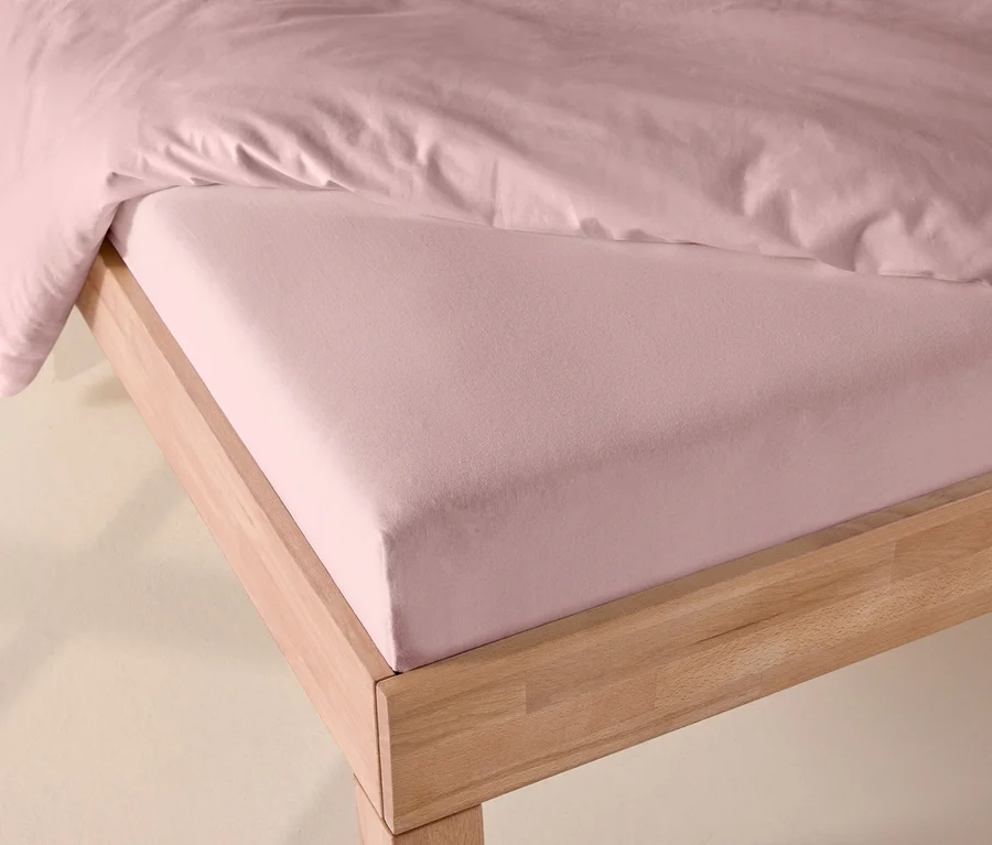 Drap-housse en jersey rose sur un lit à cadre en bois clair, partiellement recouvert d'une couette rose.
