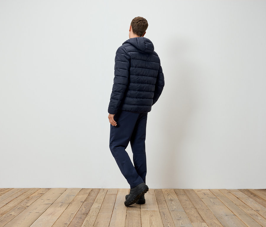 Un homme vu de dos se tient sur un plancher en bois, portant une veste à capuche et un pantalon bleu marine.