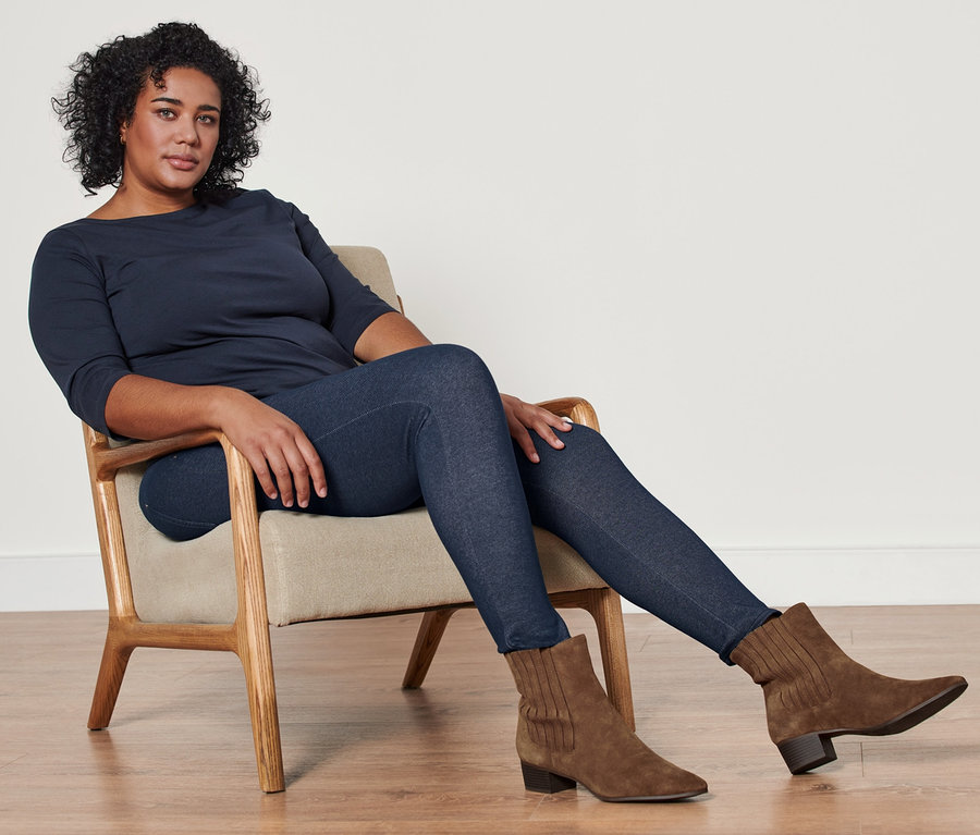 Une femme est assise dans un fauteuil et porte un t-shirt manches 3/4 bleu foncé, un legging doublé à l’aspect jean et des bottines en cuir velours.