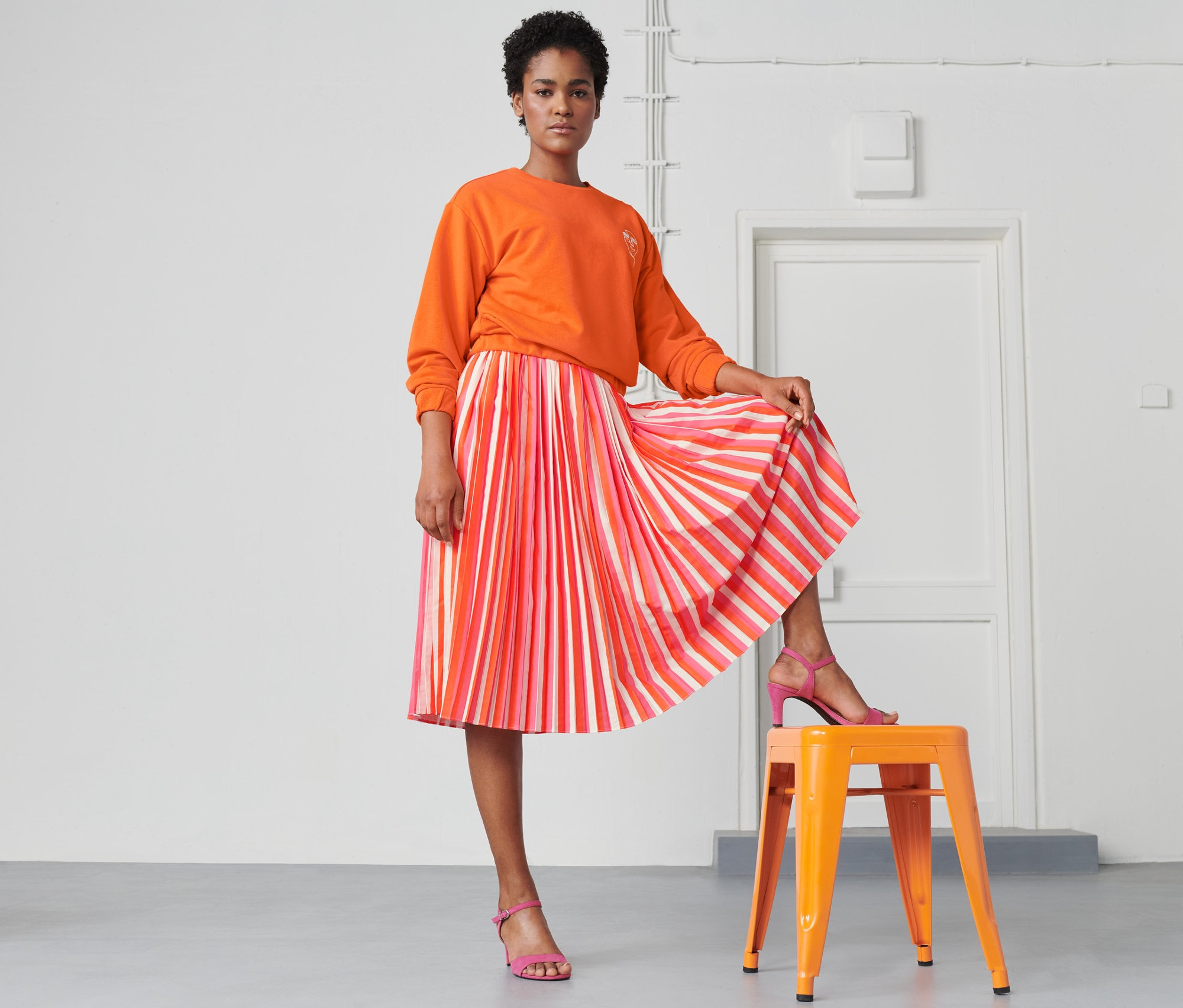 Femme posant sur un tabouret orange portant un sweat-shirt orange et une jupe plissée rayée.
