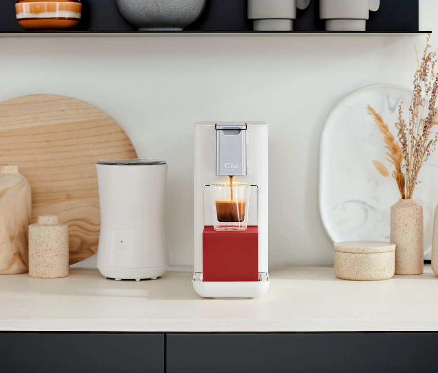 Sur un comptoir de cuisine, une machine à café Qbo prépare du café dans un verre. On voit une Qbo ICONIC Dark Chrome et une Qbo ESSENTIAL Bright Coral.