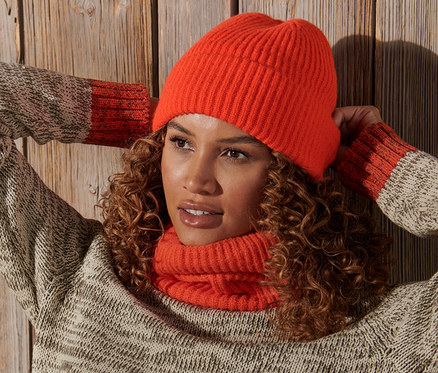 Une femme aux cheveux bouclés pose, les mains derrière la tête, portant un bonnet en maille orange.