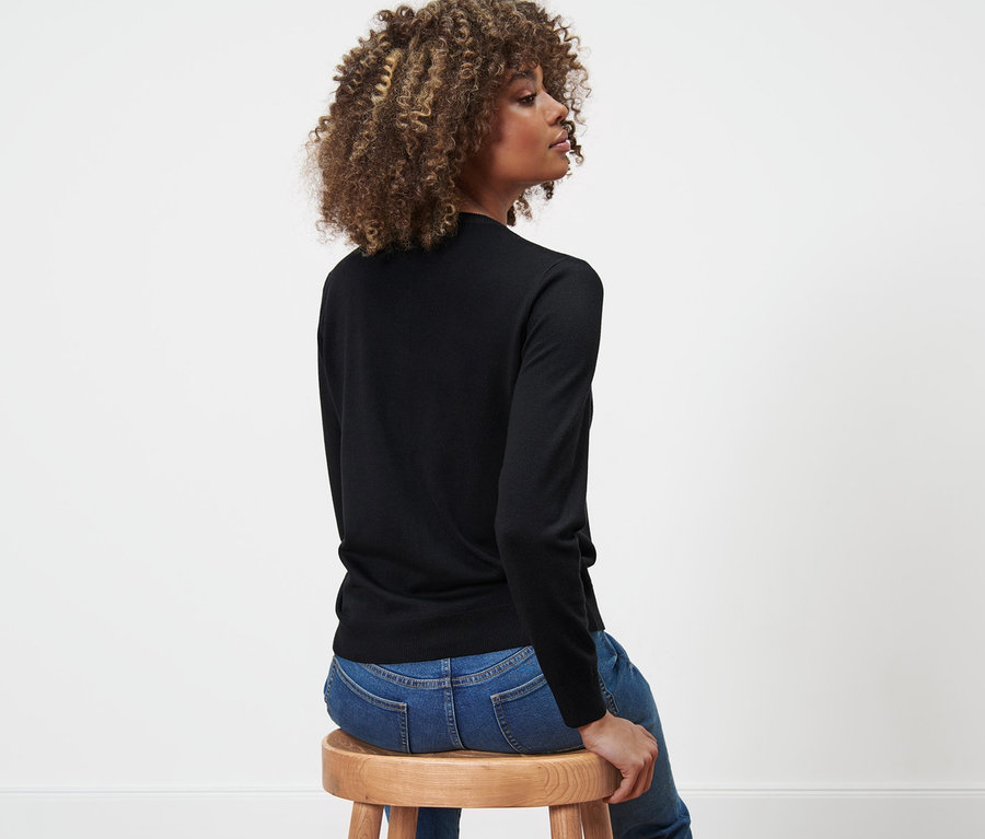 Une femme assise de côté sur un tabouret en bois, portant un cardigan en maille fine et un jean.