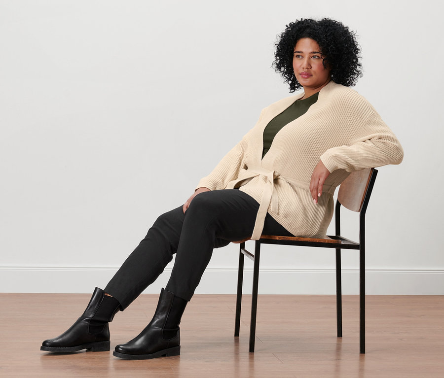 Une femme est assise sur une chaise et porte un cardigan cache-cœur, un t-shirt manches 3/4 vert olive, un pantalon stretch noir et des bottines en cuir.