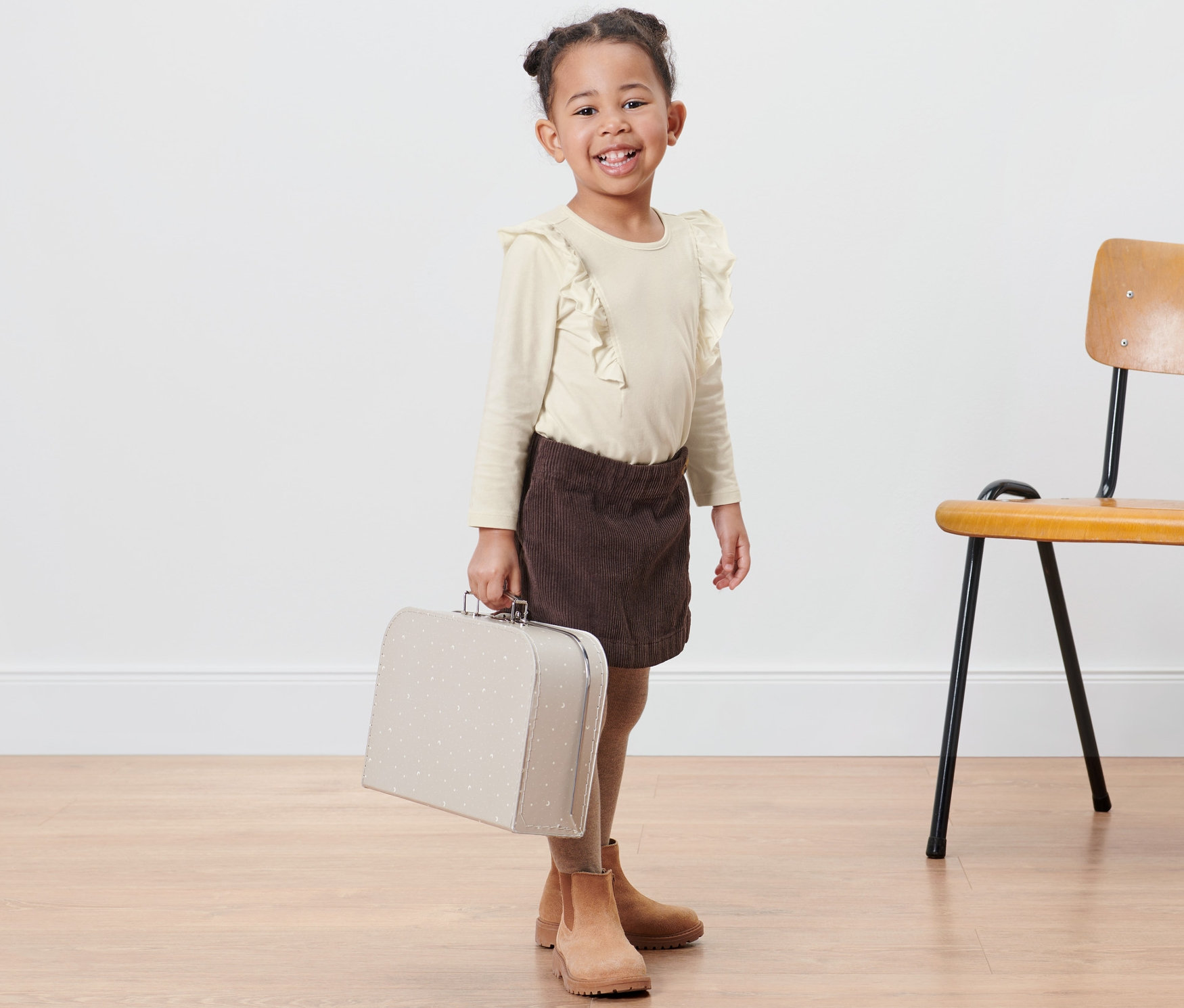 Une petite fille souriante tient une valise crème. Elle porte un t-shirt manches longues crème pour enfants et une jupe marron. Une chaise se trouve à côté d'elle.
