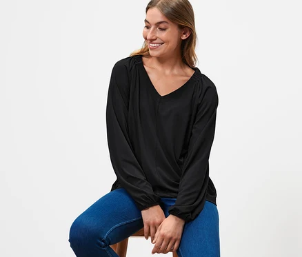 Une femme est assise sur un tabouret et porte une blouse noire avec des fronces et un jean bleu.